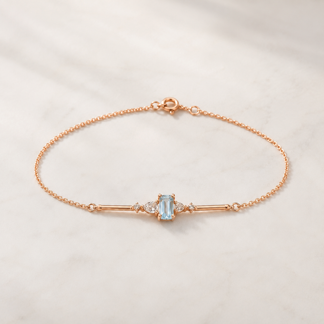 A delicate rose gold chain bracelet with a central bar setting featuring a light blue emerald-cut gemstone and small clear accent stones, displayed on a light marble surface.
