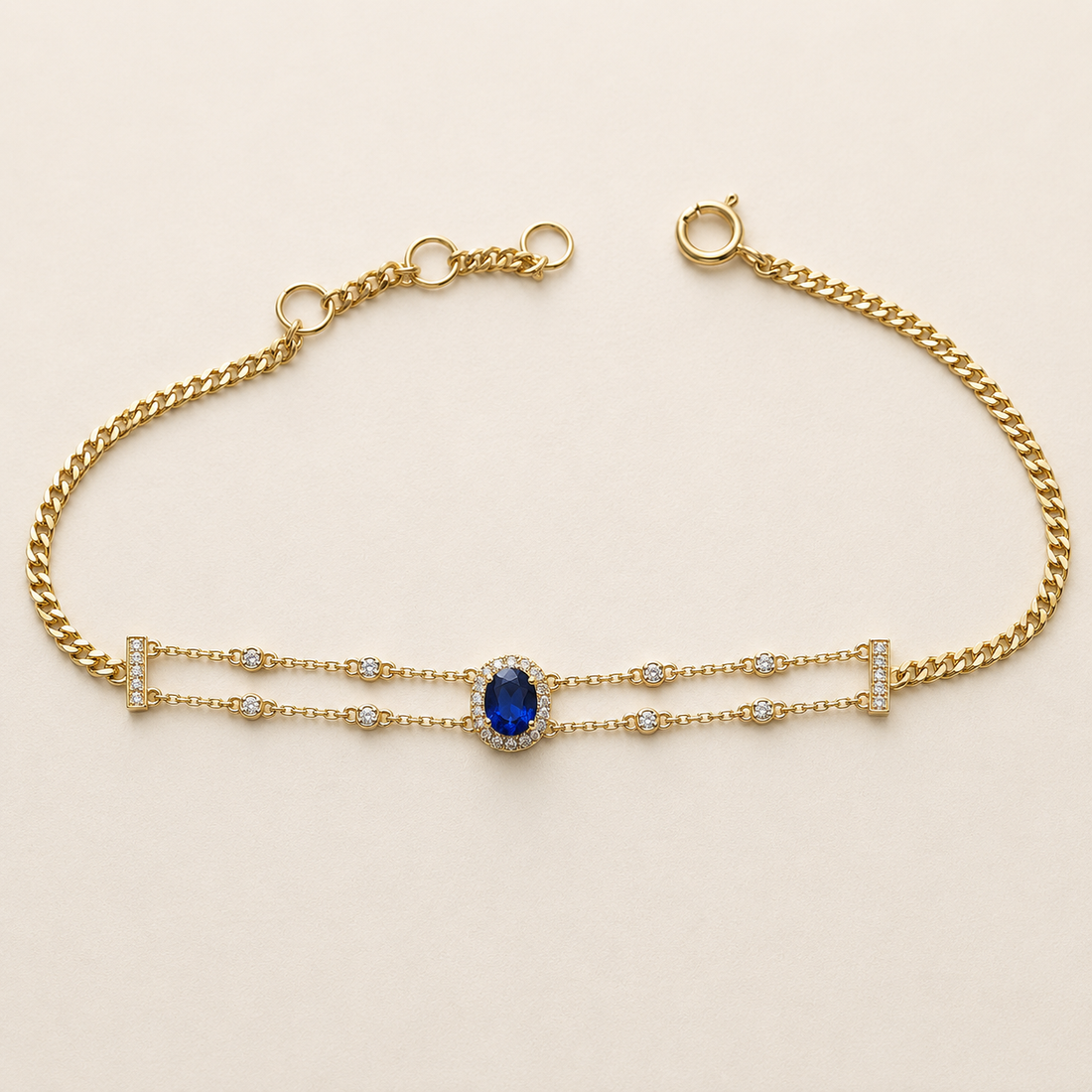 A gold chain bracelet featuring a central oval blue gemstone surrounded by small clear stones, with delicate gold links extending from the sides, photographed on a light beige background.