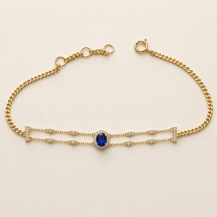 A gold chain bracelet featuring a central oval blue gemstone surrounded by small clear stones, with delicate gold links extending from the sides, photographed on a light beige background.