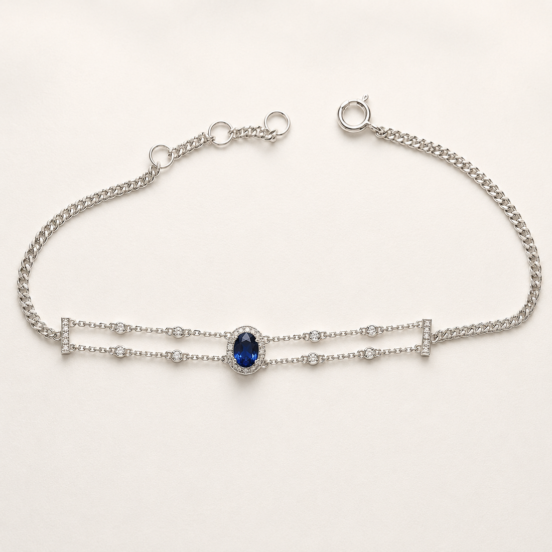 A silver double-strand chain bracelet featuring a central oval-cut deep blue gemstone surrounded by small clear stones, displayed on a clean white background.