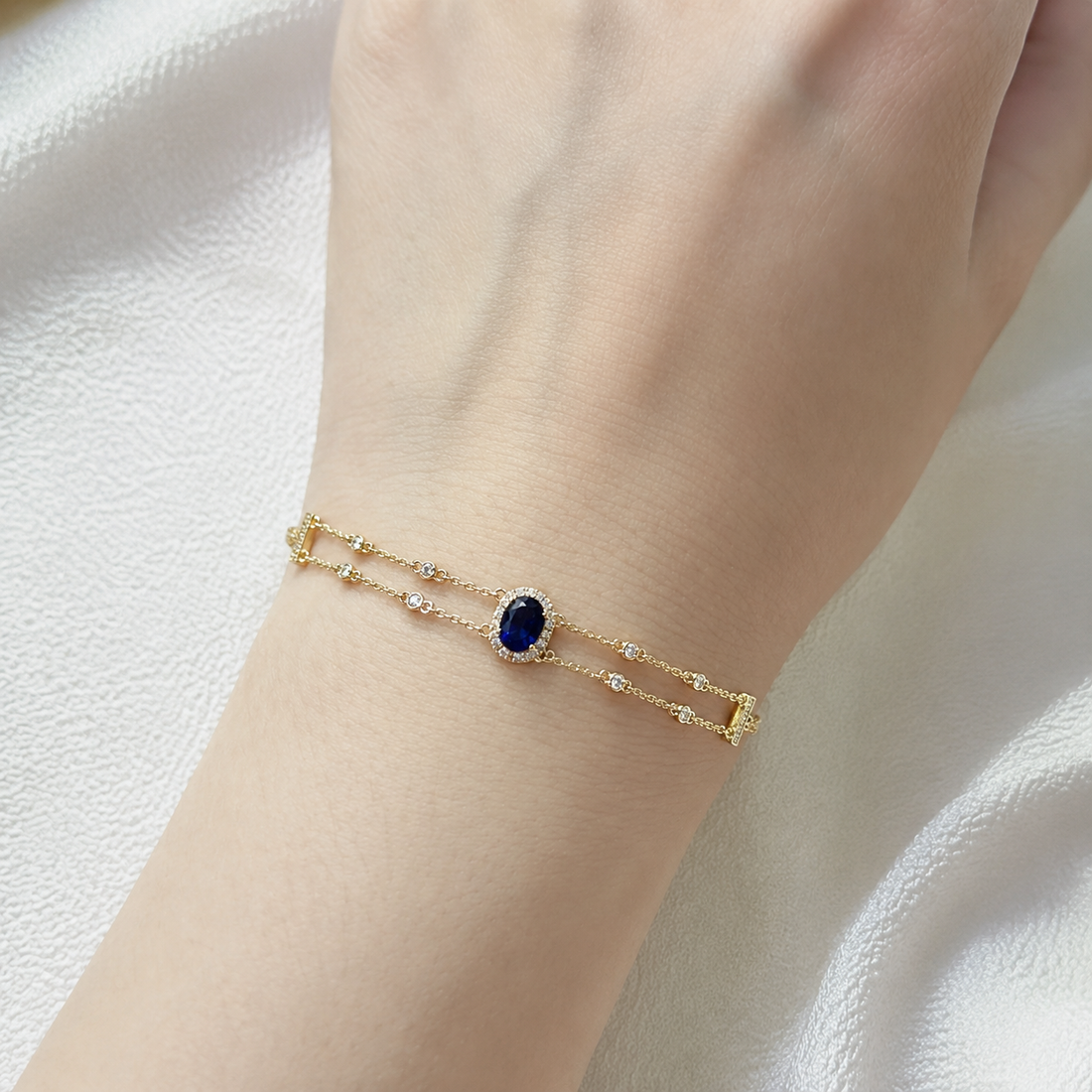 A delicate gold double-strand bracelet with a central oval-cut dark blue gemstone surrounded by small clear stones, shown worn on a person's wrist against a soft white fabric background.