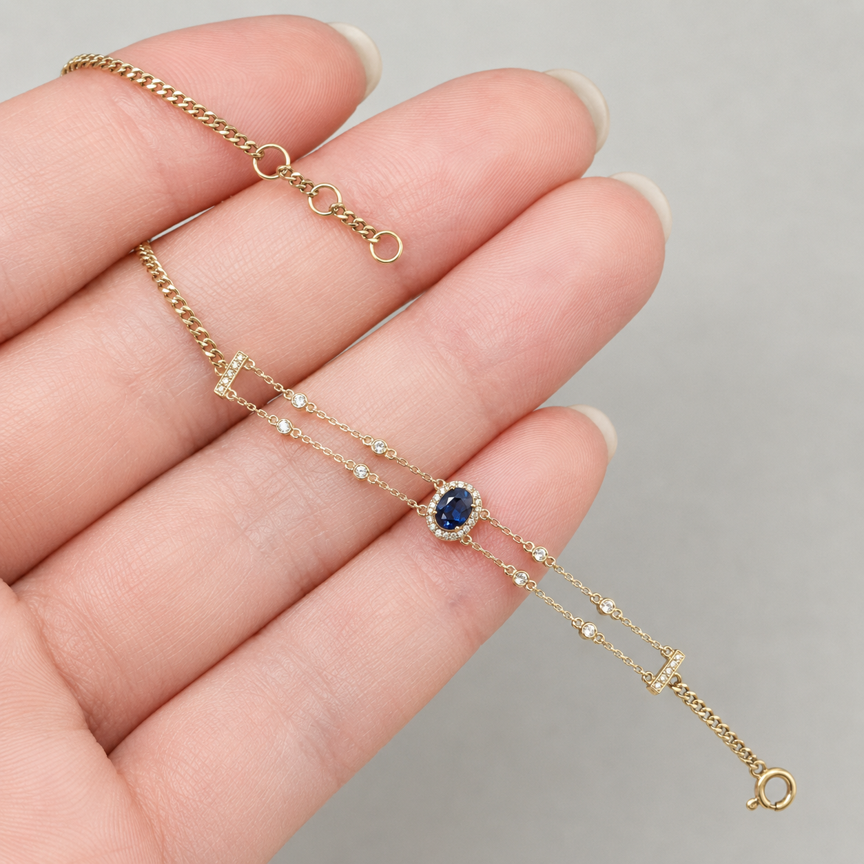 A delicate gold chain bracelet with a central oval blue gemstone surrounded by small clear stones, displayed on a person's hand against a neutral gray background.