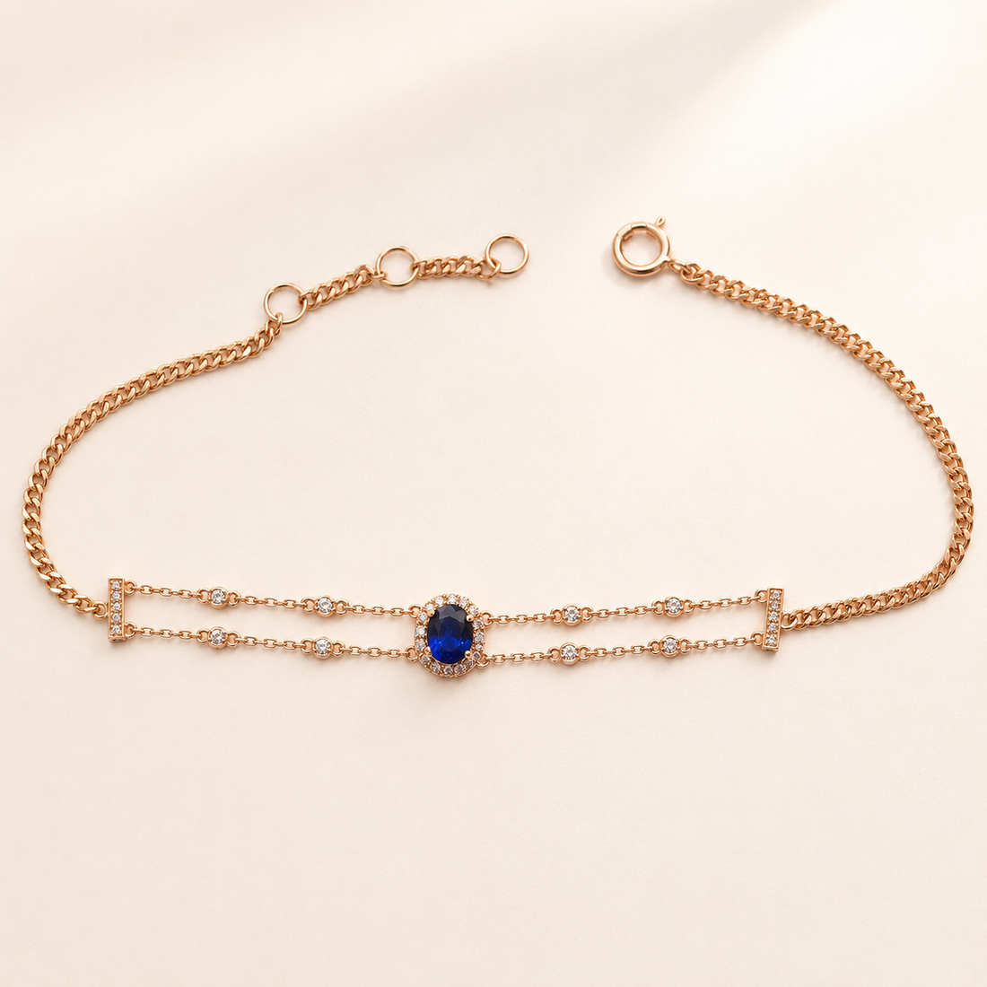 A delicate rose gold chain bracelet featuring a central oval blue gemstone surrounded by small clear stones, displayed on a light cream background.