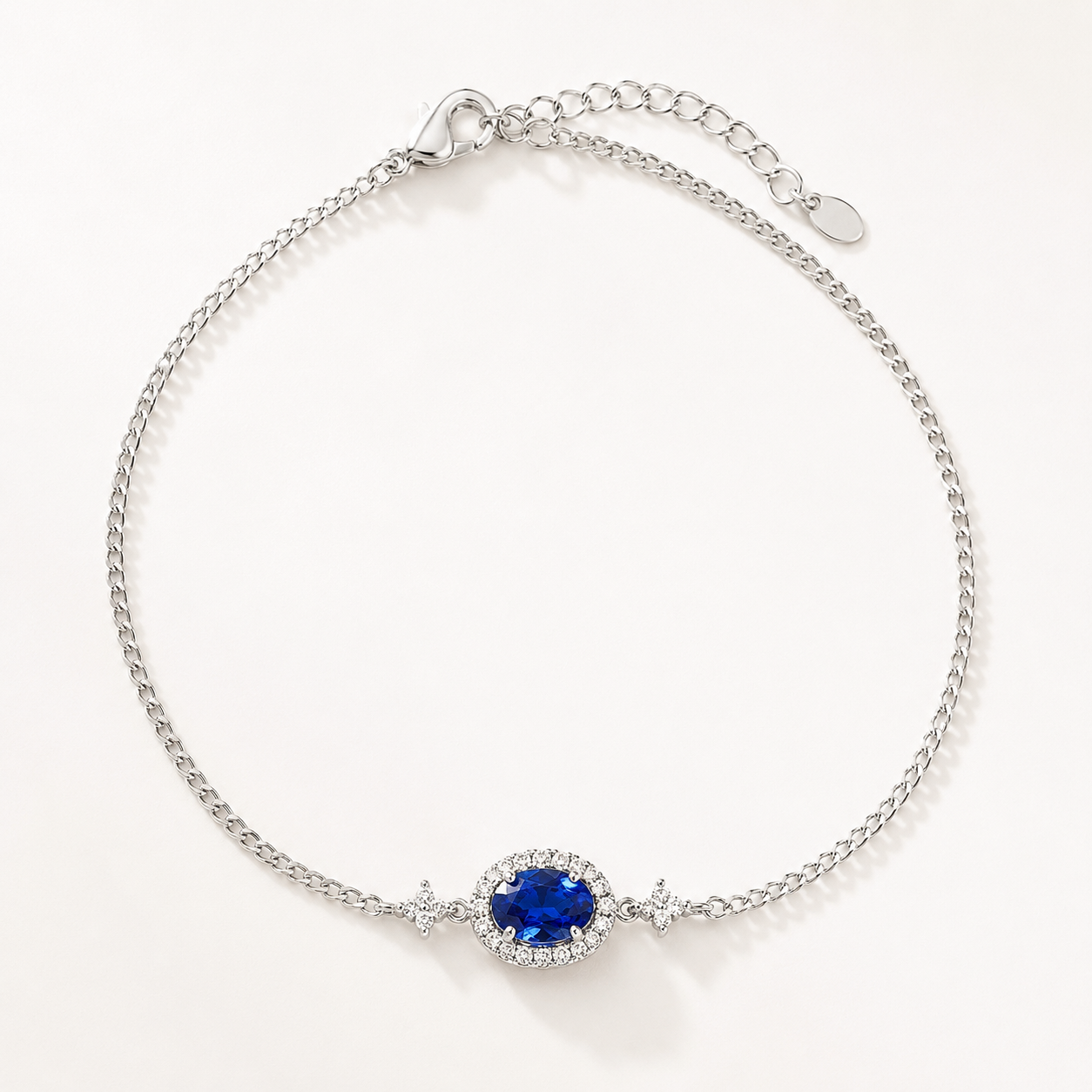 A silver chain bracelet with a central oval blue gemstone surrounded by a halo of small clear stones, photographed on a clean white background.