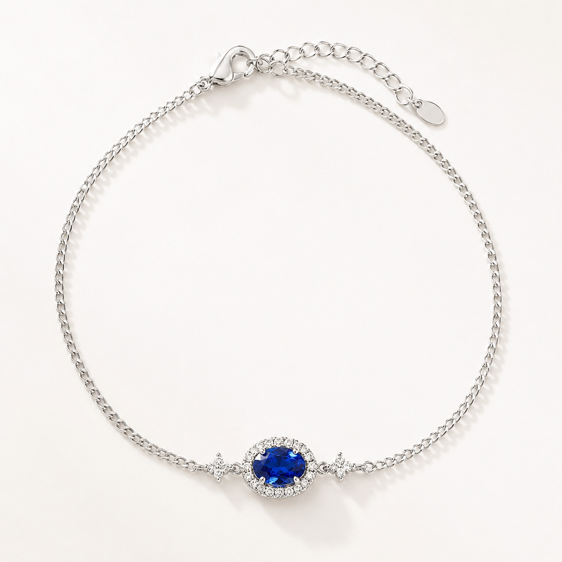 A silver chain bracelet with a central oval blue gemstone surrounded by a halo of small clear stones, photographed on a clean white background.
