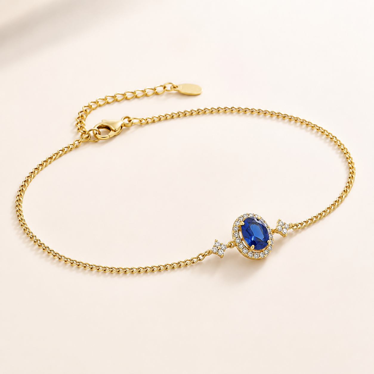 A delicate gold chain bracelet with an oval blue gemstone centerpiece, surrounded by small clear stones, displayed on a soft cream background.