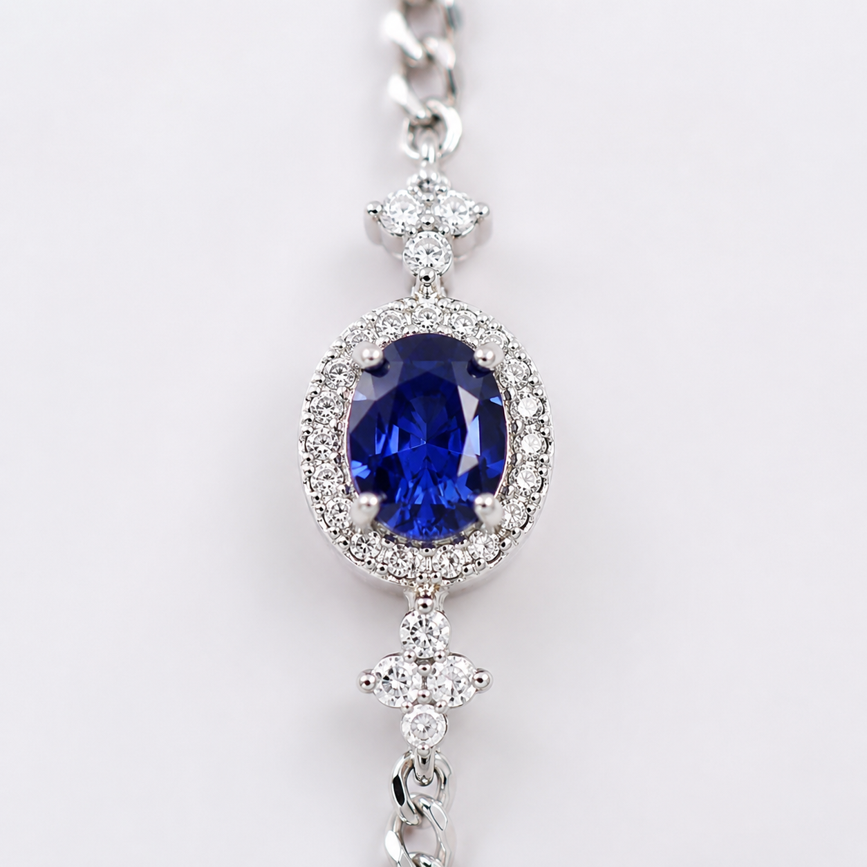 A close-up of a silver-toned jewelry piece featuring a prominent oval-cut deep blue sapphire gemstone surrounded by a halo of small clear stones, with a matching chain visible at the top and bottom.