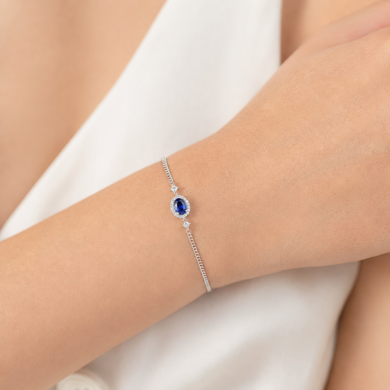 A delicate silver chain bracelet with a central oval blue gemstone and small clear accent stones, shown worn on a person's wrist against a soft white garment background.
