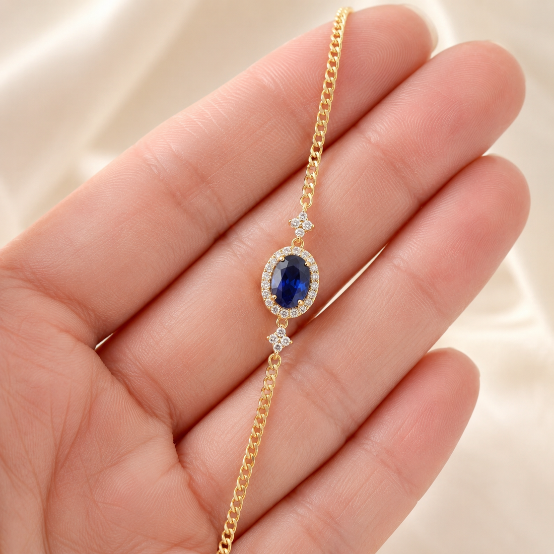 A gold chain bracelet with an oval blue gemstone surrounded by small clear stones, held in a person's hand against a soft cream background.