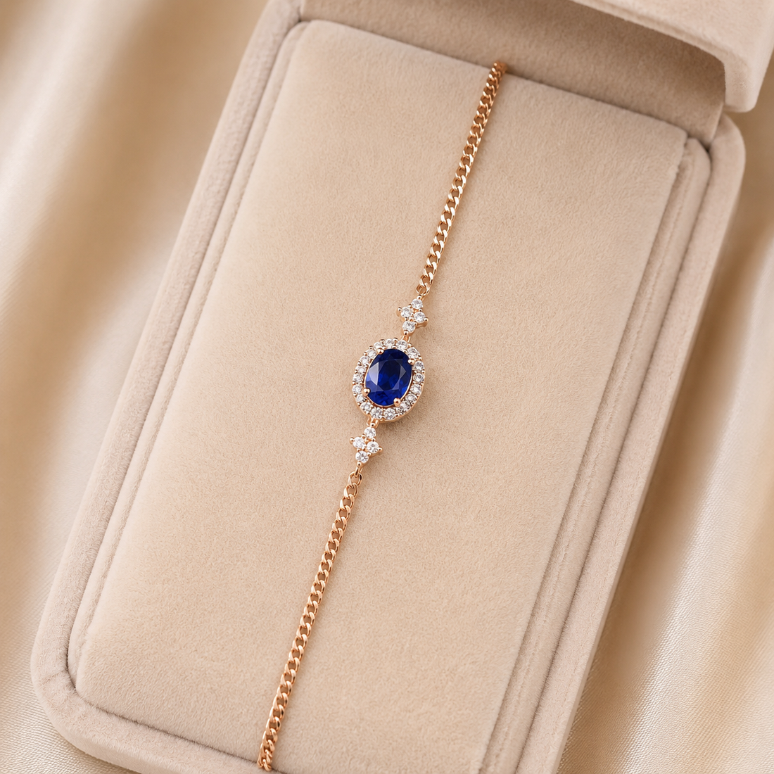 A delicate rose gold chain bracelet with an oval blue gemstone surrounded by small clear stones, displayed on a beige jewelry box.
