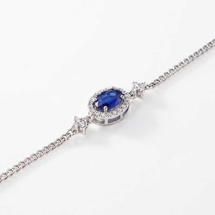 A silver-toned chain bracelet featuring an oval centerpiece with a deep blue gemstone surrounded by small clear stones, set against a clean white background.