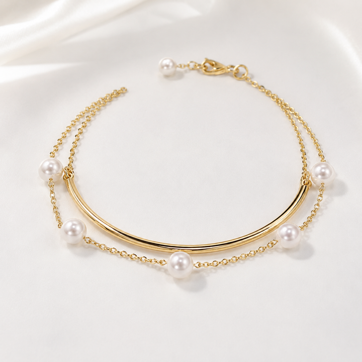 A gold-toned double-layered chain anklet featuring a curved center bar and multiple white pearl beads, displayed on a soft white satin fabric background.