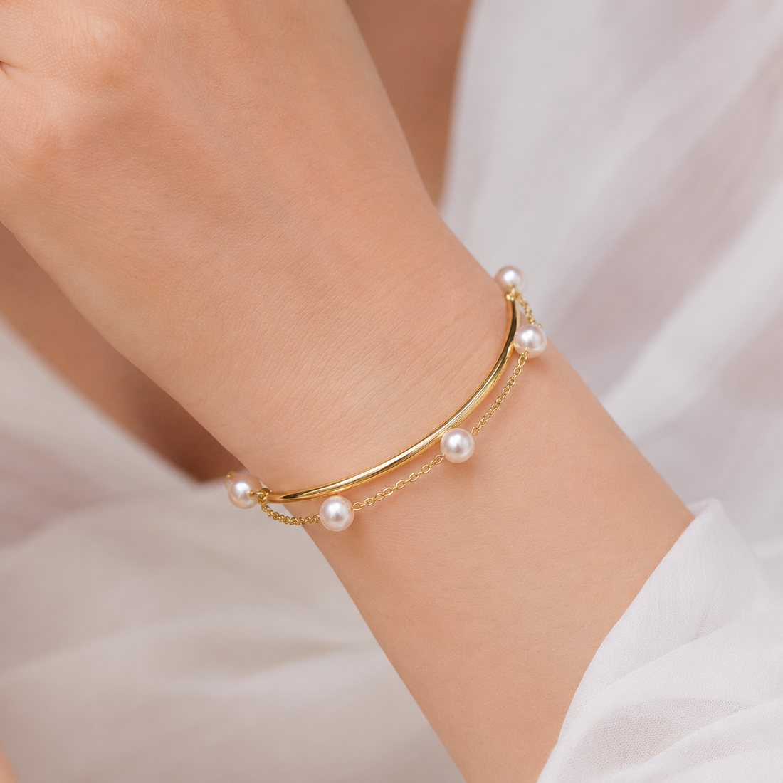 A delicate gold bangle bracelet with small white pearls, worn on a person's wrist against a soft white fabric background.