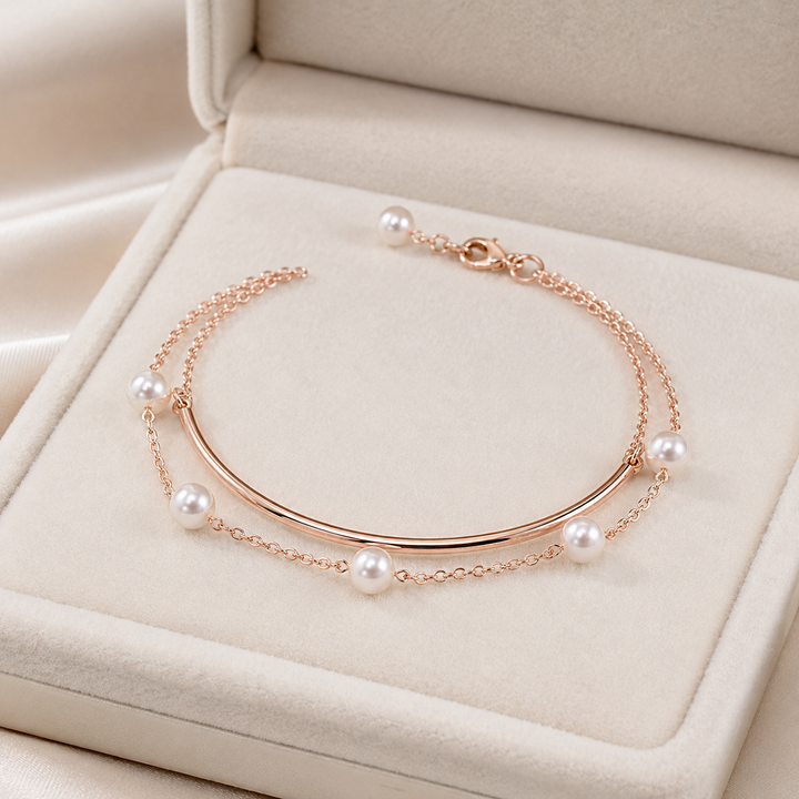 A rose gold double-strand bangle bracelet with white pearl accents, displayed inside an open cream-colored jewelry box.
