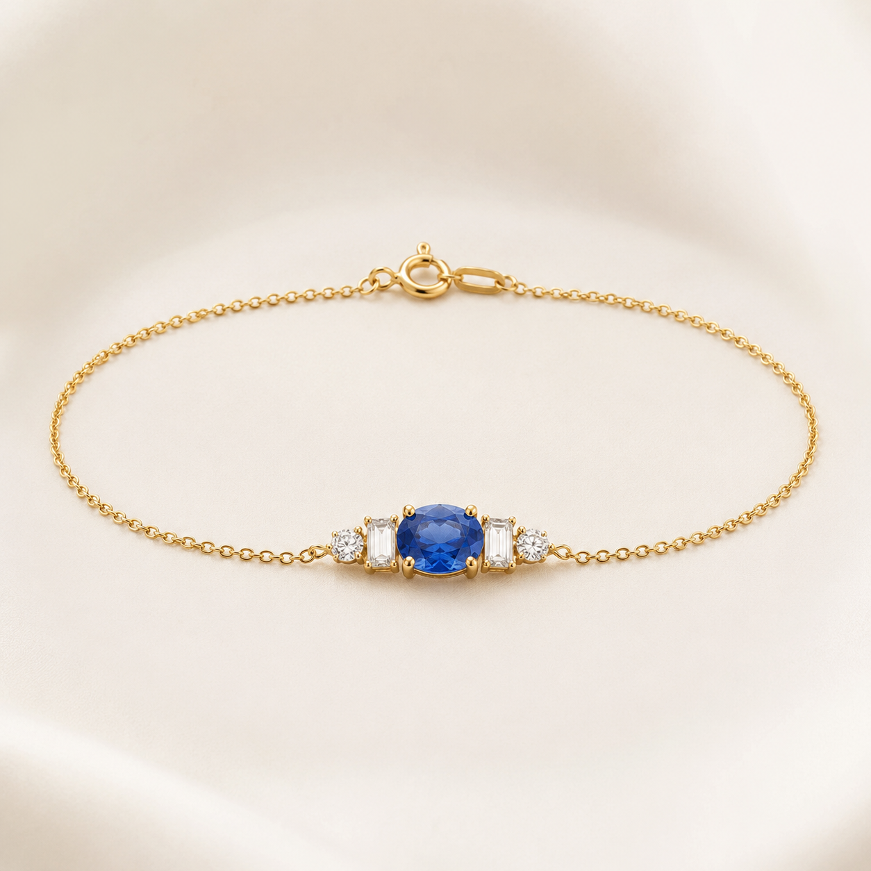 A delicate gold chain bracelet featuring a central oval blue gemstone flanked by two small clear stones, displayed on a soft cream-colored background.