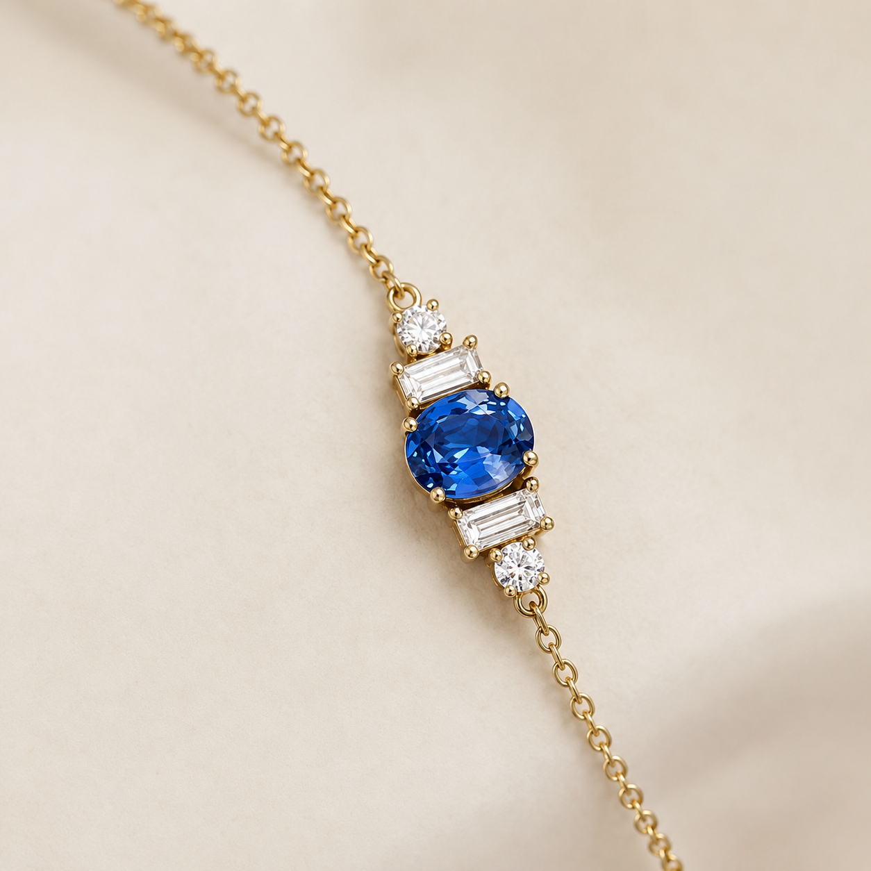 A delicate gold chain bracelet featuring a central cluster with a round blue sapphire and baguette-cut clear stones, photographed on a soft beige background.
