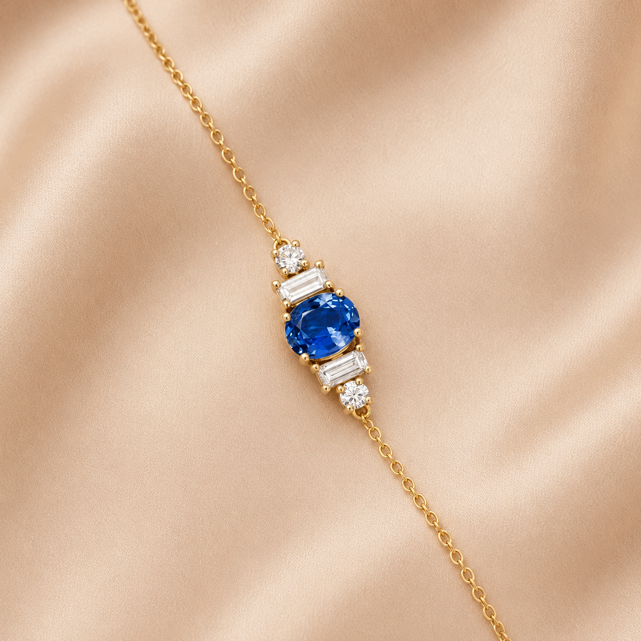 A delicate yellow gold chain bracelet featuring a central cluster with a round-cut blue sapphire and baguette-cut clear stones, displayed on a soft beige satin fabric background.