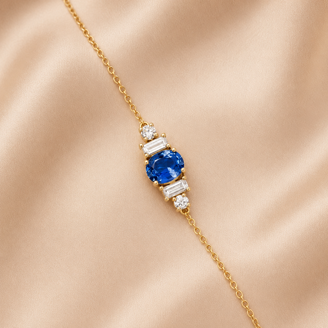 A delicate yellow gold chain bracelet featuring a central cluster with a round-cut blue sapphire and baguette-cut clear stones, displayed on a soft beige satin fabric background.