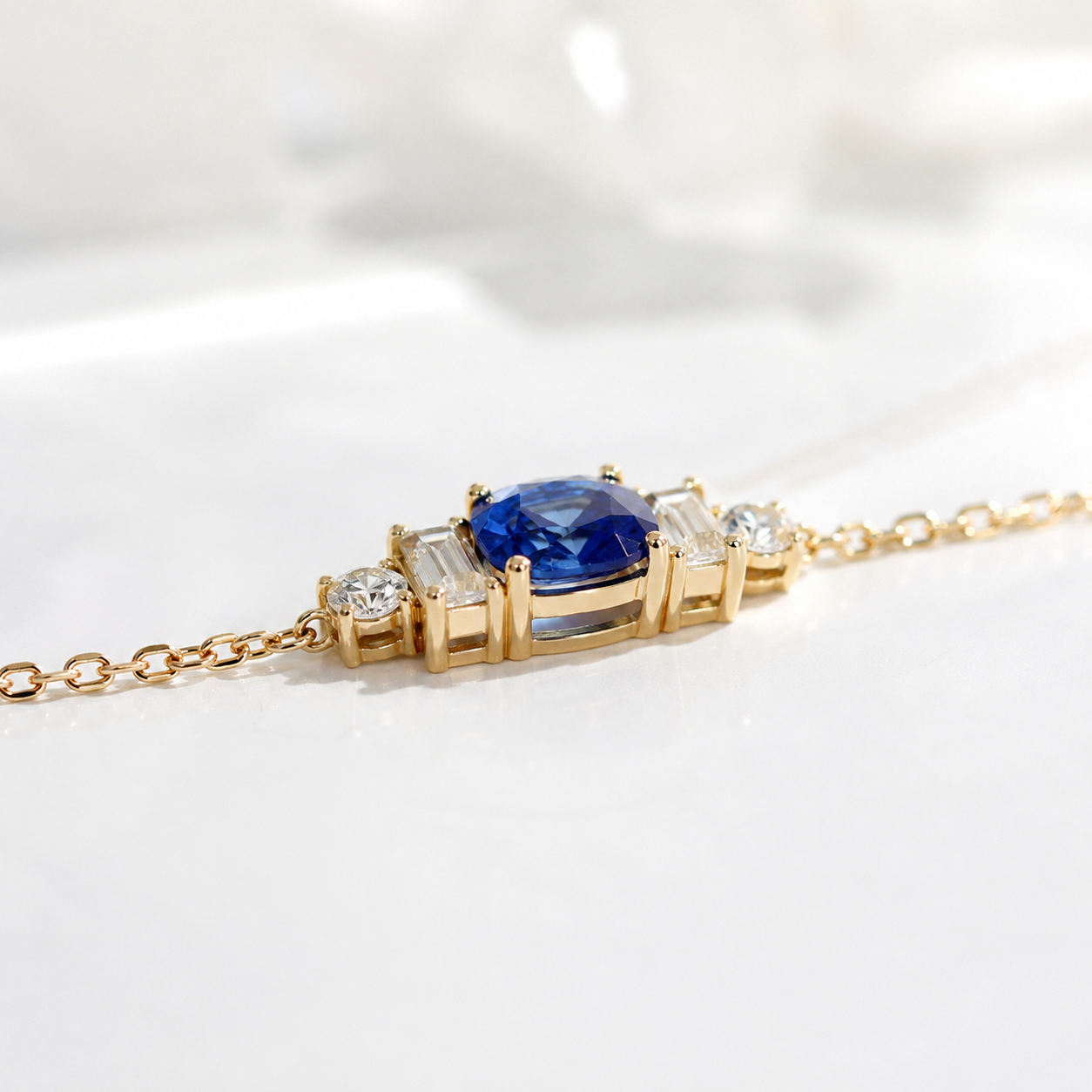 A gold chain necklace featuring a rectangular centerpiece with a prominent blue cushion-cut gemstone, flanked by baguette-cut and round clear stones, set against a soft white background.