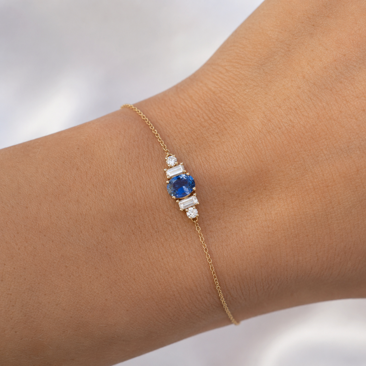A delicate gold chain bracelet with a central blue sapphire and clear baguette and round stones, shown worn on a person's wrist.