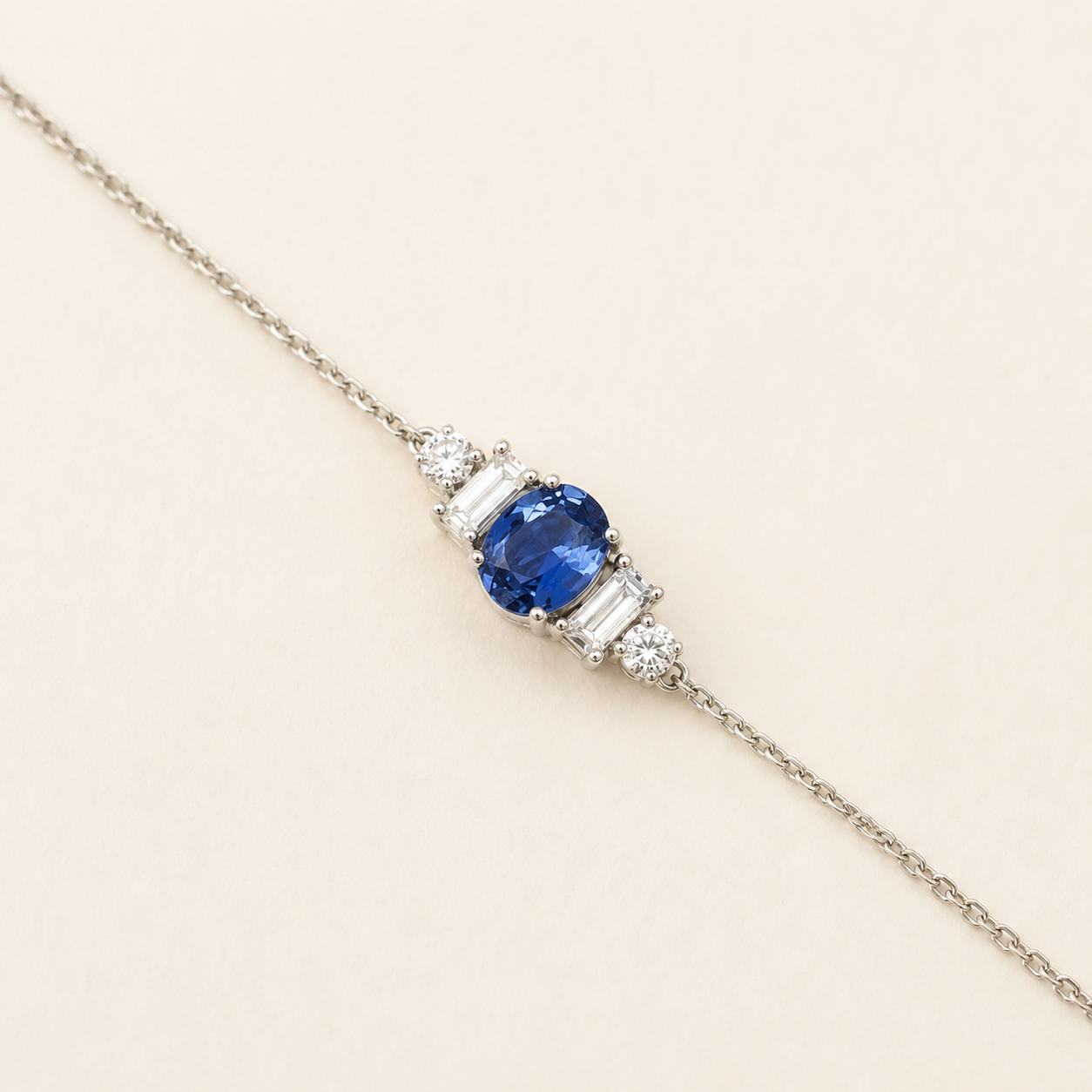 A delicate silver chain bracelet featuring a central oval-cut blue sapphire gemstone flanked by baguette-cut and round-cut clear stones, set against a solid light beige background.