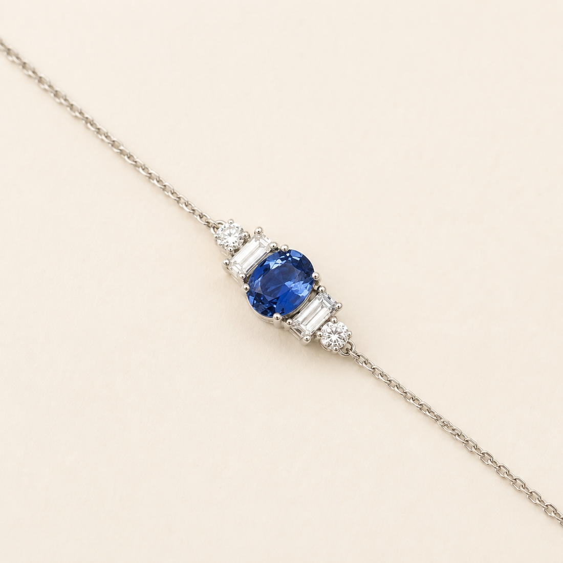 A delicate silver chain bracelet featuring a central oval-cut blue sapphire gemstone flanked by baguette-cut and round-cut clear stones, set against a solid light beige background.
