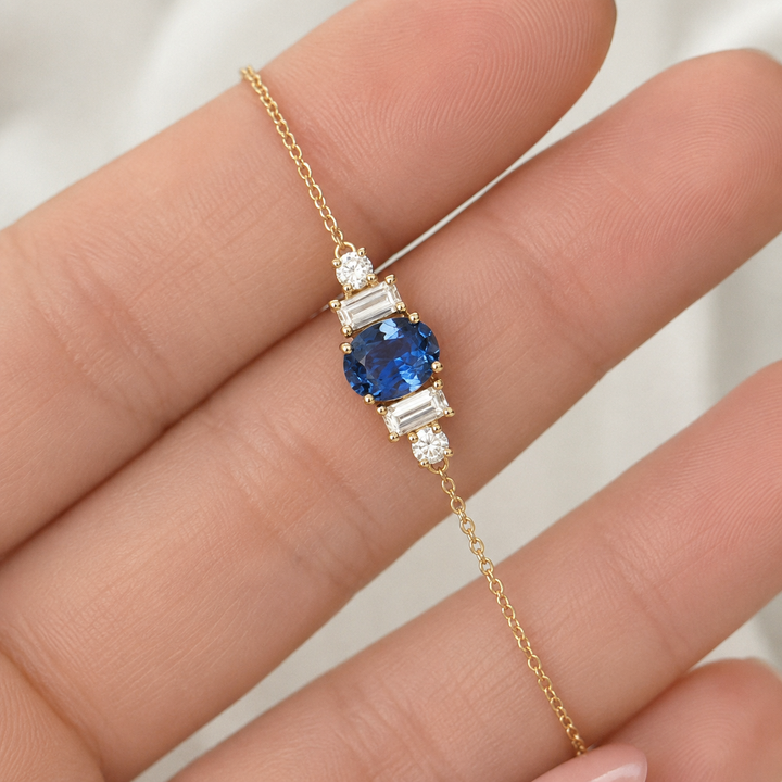 A delicate gold chain bracelet with a central cluster featuring a round blue sapphire and baguette-cut clear stones, shown resting on a person's fingers.