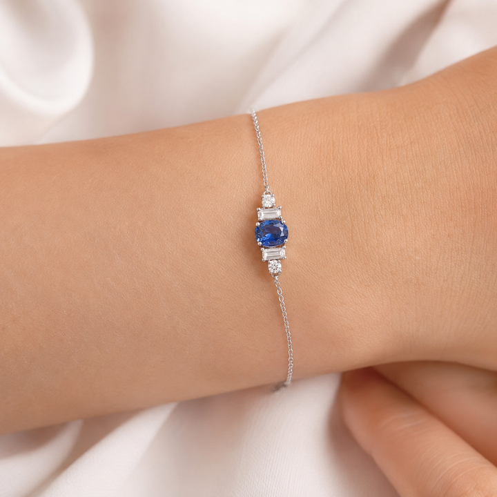 A delicate silver chain bracelet with a central rectangular blue sapphire and small clear accent stones, shown worn on a person's wrist against a soft white fabric background.