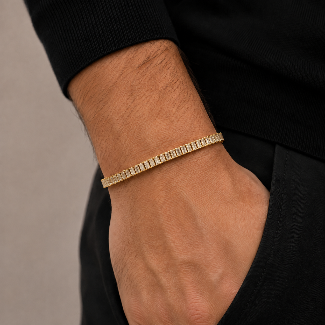 A gold tennis bracelet with baguette-cut stones worn on a man's wrist, shown in a close-up lifestyle shot against a black shirt and dark background.