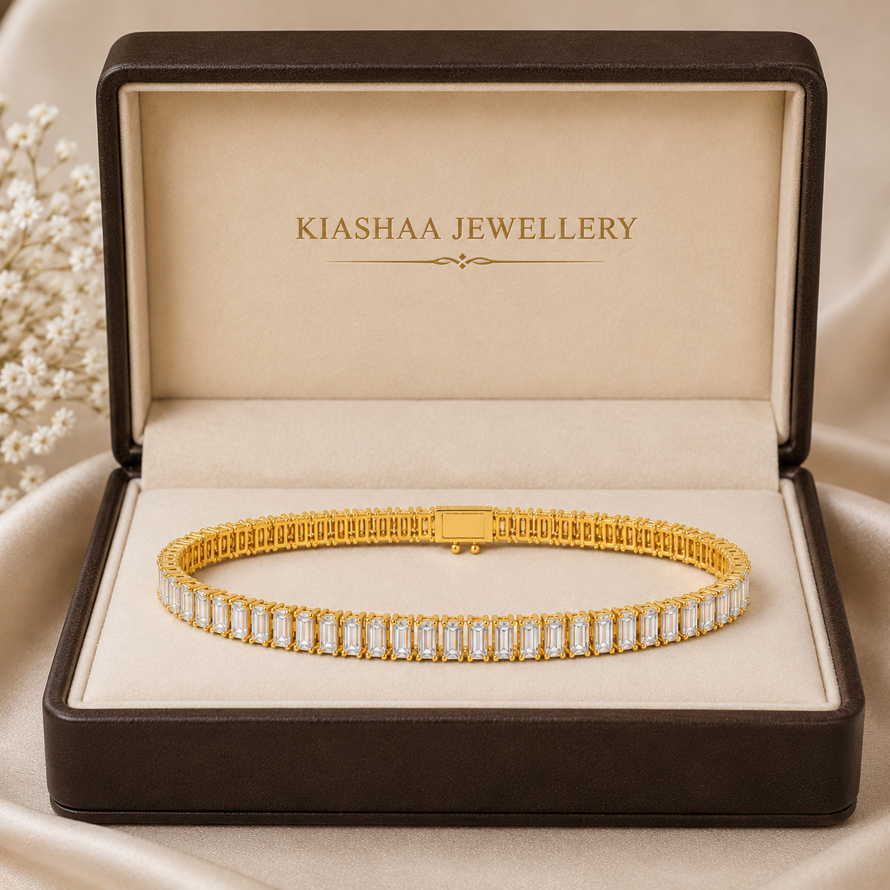A gold tennis bracelet with rectangular clear stones, displayed inside an open brown jewelry box with the brand name 'KIASHAA JEWELLERY' on the inner lid, set against a soft beige satin background.