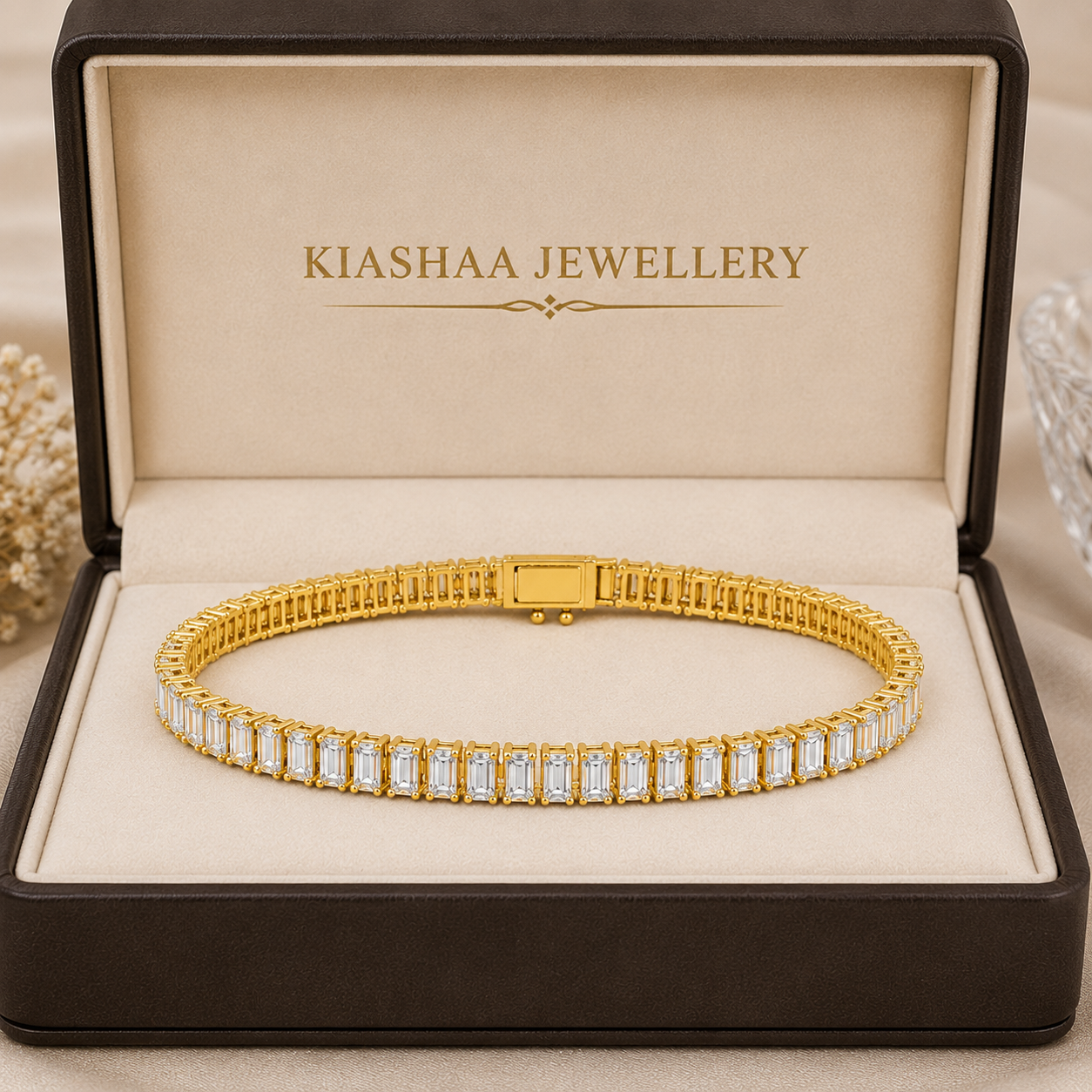 A gold tennis bracelet with rectangular clear stones, presented inside an open dark brown jewelry box with a beige interior. The inside lid is printed with the brand name 'KIASHAA JEWELLERY'.
