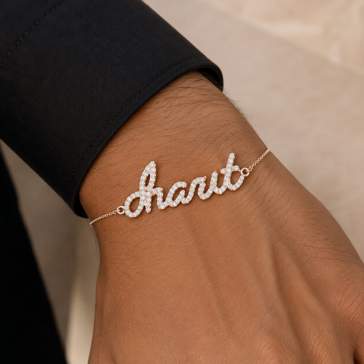 A close-up of a person's wrist wearing a delicate nameplate bracelet in cursive script spelling charit with sparkling clear stones, set against a black sleeve and a soft beige background.