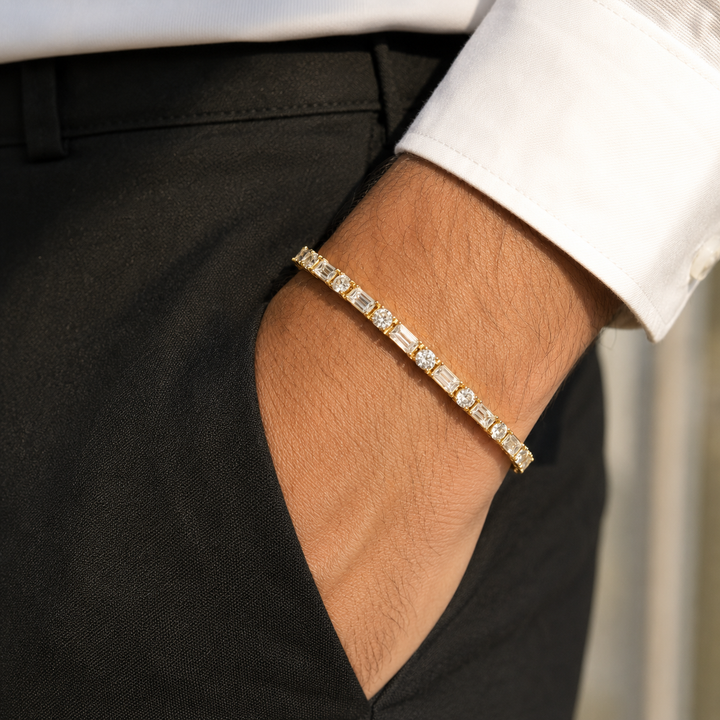 A gold tennis bracelet with baguette and round-cut clear stones worn on a man's wrist, shown with a white dress shirt cuff and black trousers.