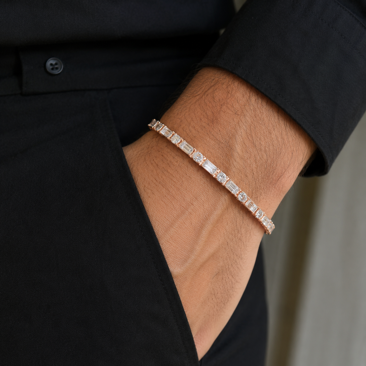 A close-up of a man's wrist wearing a rose gold tennis bracelet with baguette and round-cut stones, styled with a black dress shirt.