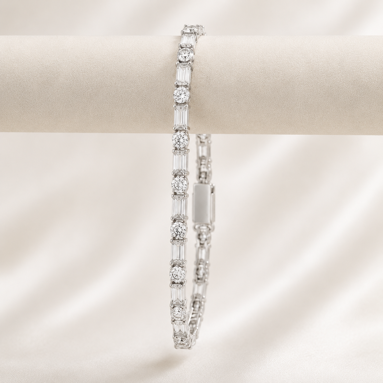A silver tennis bracelet with round and baguette-cut clear stones, displayed on a cream-colored jewelry stand against a soft, draped satin background.