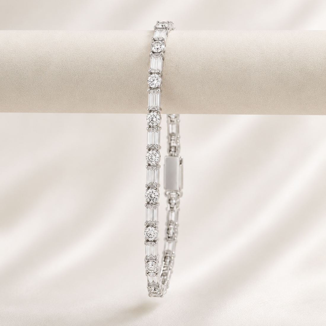 A silver tennis bracelet with round and baguette-cut clear stones, displayed on a cream-colored jewelry stand against a soft, draped satin background.