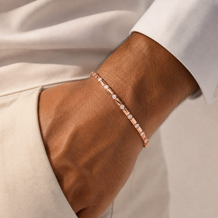 A close-up of a man's wrist wearing a delicate rose gold bracelet with small clear stones, styled with a white shirt and beige trousers.