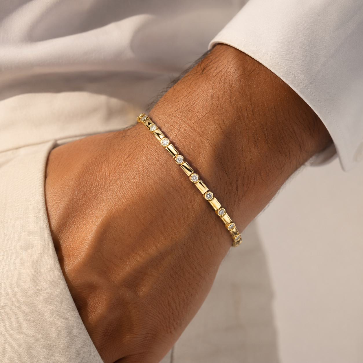 A close-up of a gold tennis bracelet with small clear stones, worn on a person's wrist. The person is wearing a white collared shirt and light beige trousers.