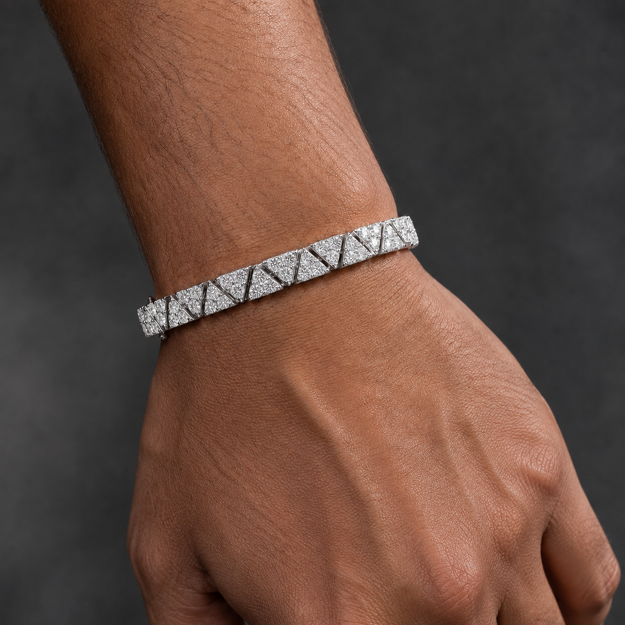 A close-up of a person's wrist and hand wearing a silver-toned tennis bracelet with a repeating triangular pattern of pavé-set clear stones, set against a dark gray background.