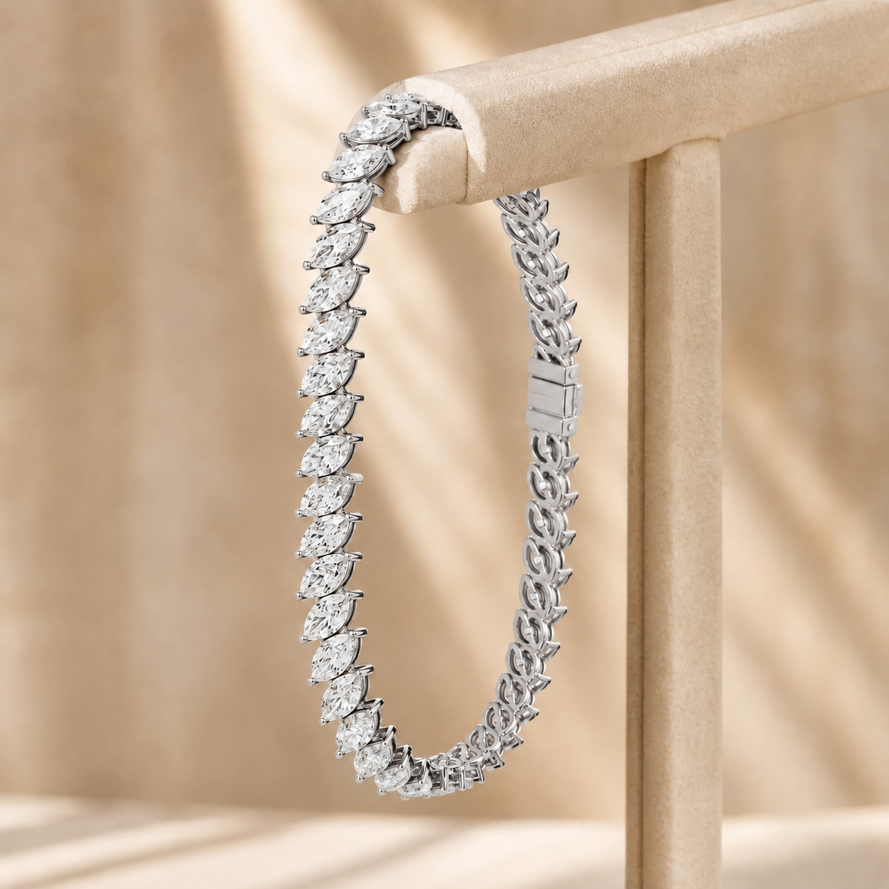 A silver tennis bracelet with marquise-cut gemstones, draped over a beige jewelry display stand against a warm beige background with soft shadows.