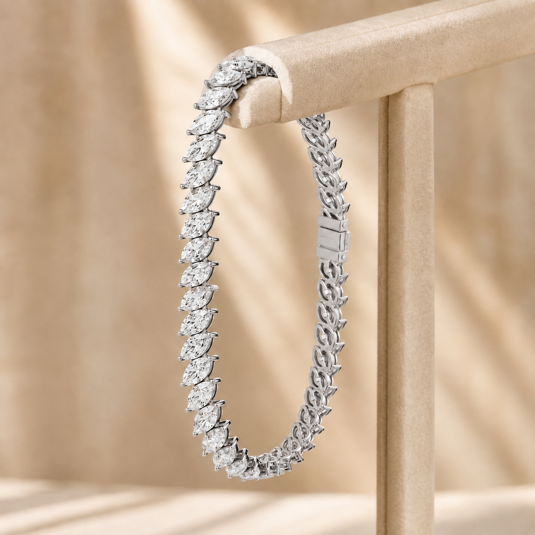 A silver tennis bracelet with marquise-cut gemstones, draped over a beige jewelry display stand against a warm beige background with soft shadows.