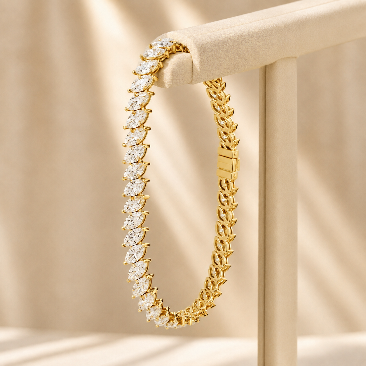 A gold tennis bracelet with marquise-cut clear stones, displayed on a beige jewelry stand against a softly lit beige fabric background.