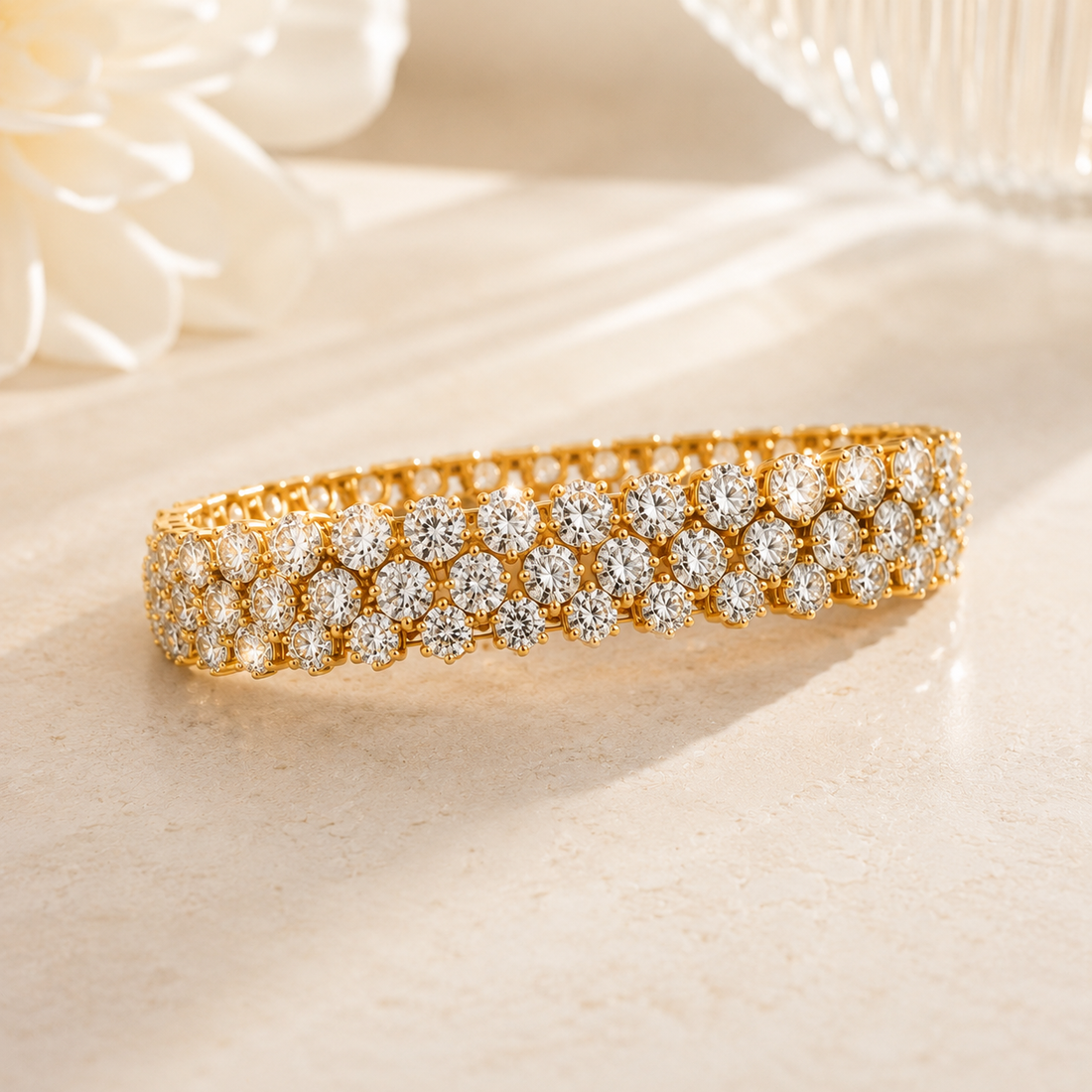 A gold tennis bracelet adorned with rows of sparkling clear gemstones, displayed on a light beige surface with soft-focus white floral elements in the background.