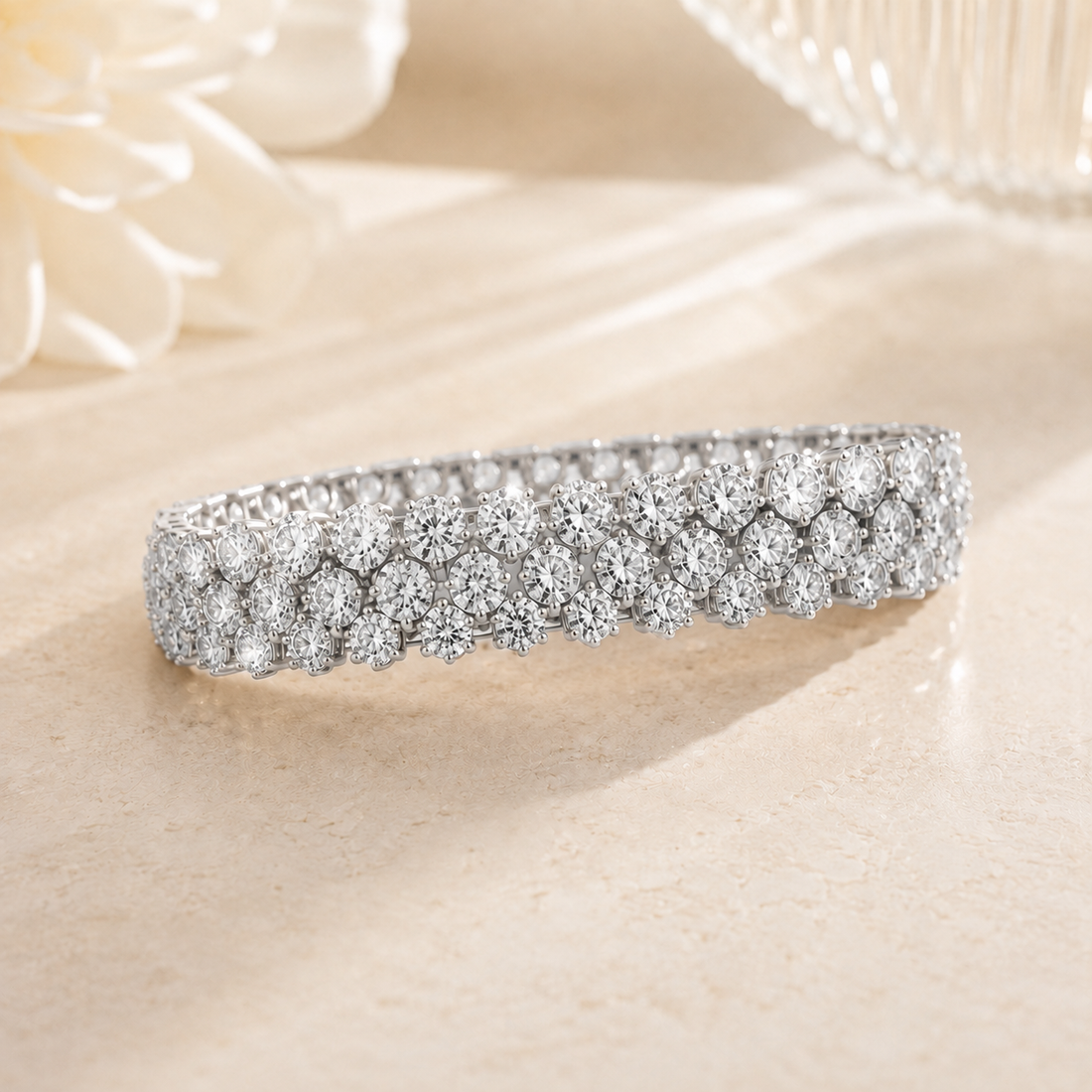 A silver-toned diamond tennis bracelet with a double row of round-cut diamonds, displayed on a light beige surface with soft-focus decorative flowers in the background.