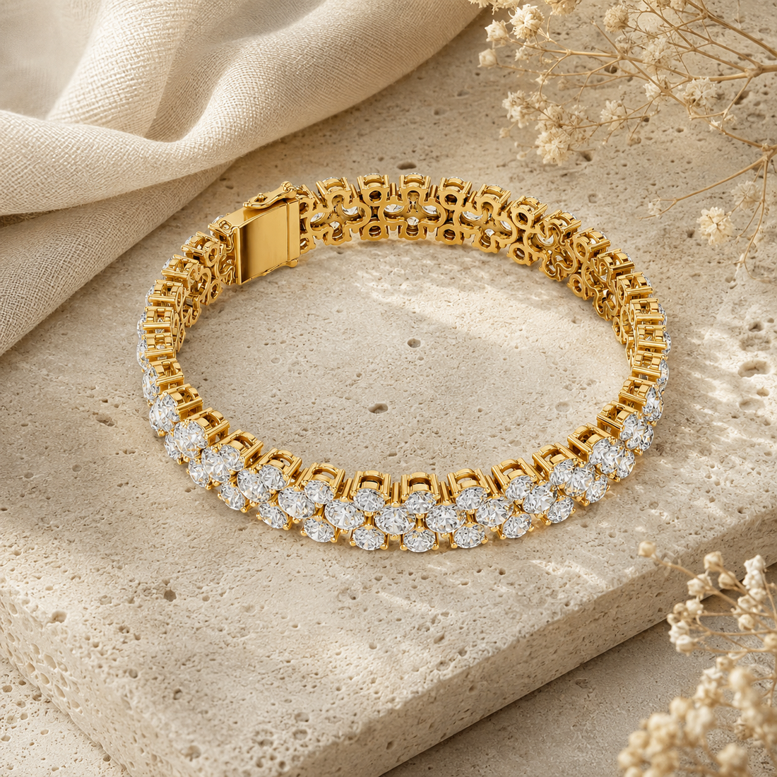 A gold tennis bracelet adorned with sparkling clear gemstones, displayed on a textured beige stone slab with draped beige fabric and dried floral accents.