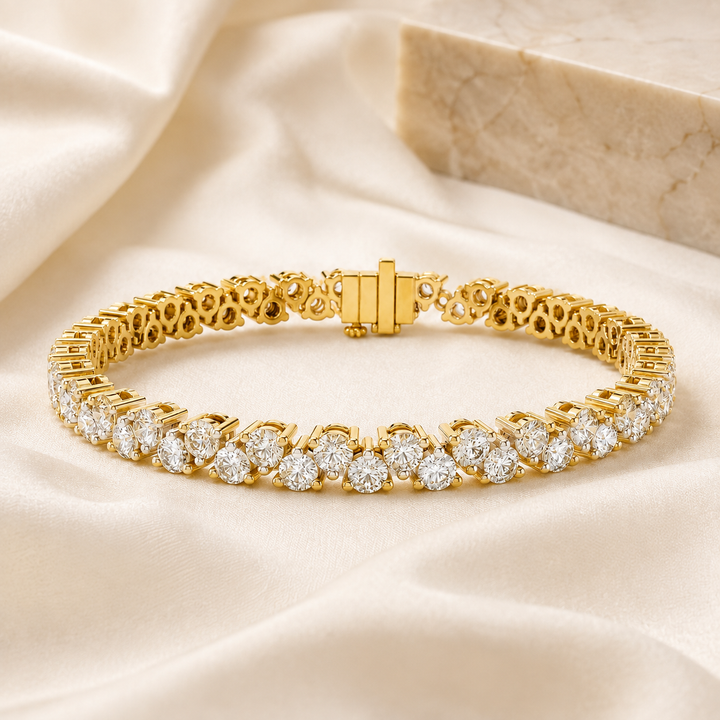 A gold tennis bracelet with round-cut clear stones, displayed on draped cream satin fabric with a marble block in the background.