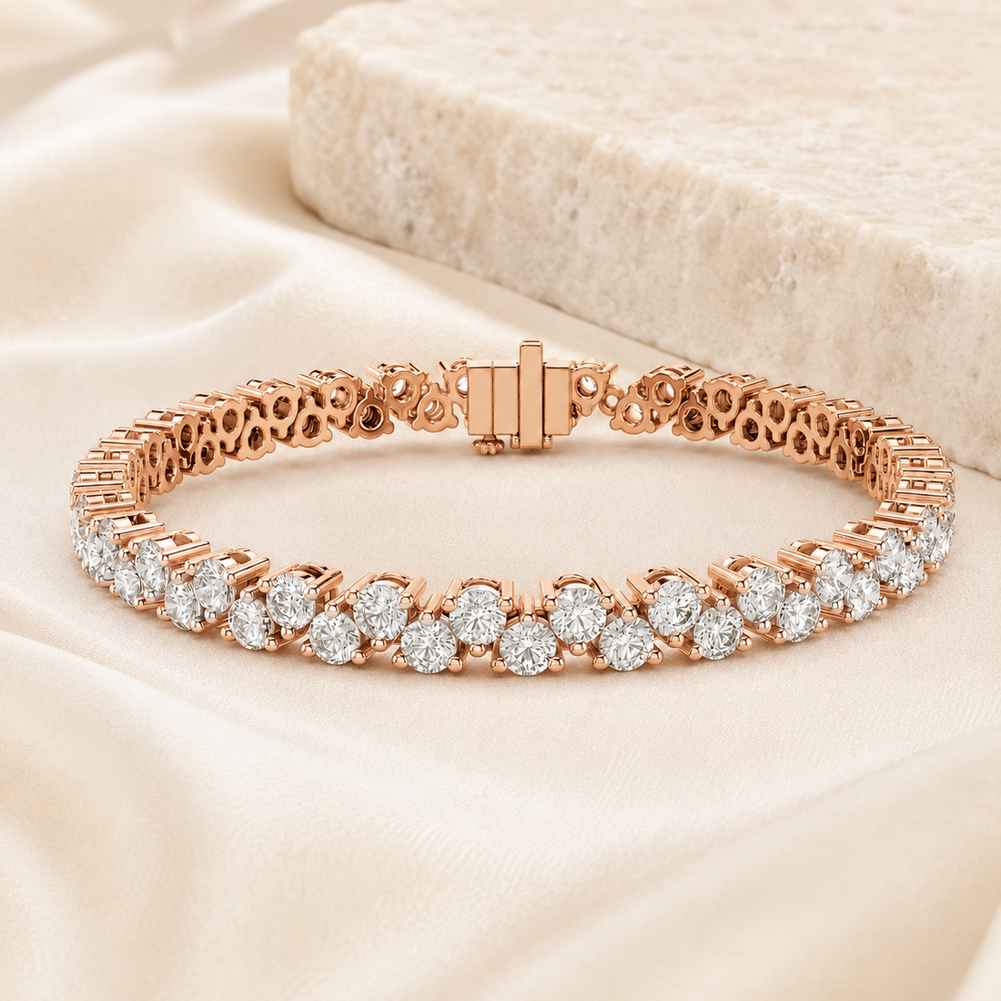 A rose gold tennis bracelet with round-cut clear stones, displayed on draped cream satin fabric with a textured stone prop in the background.