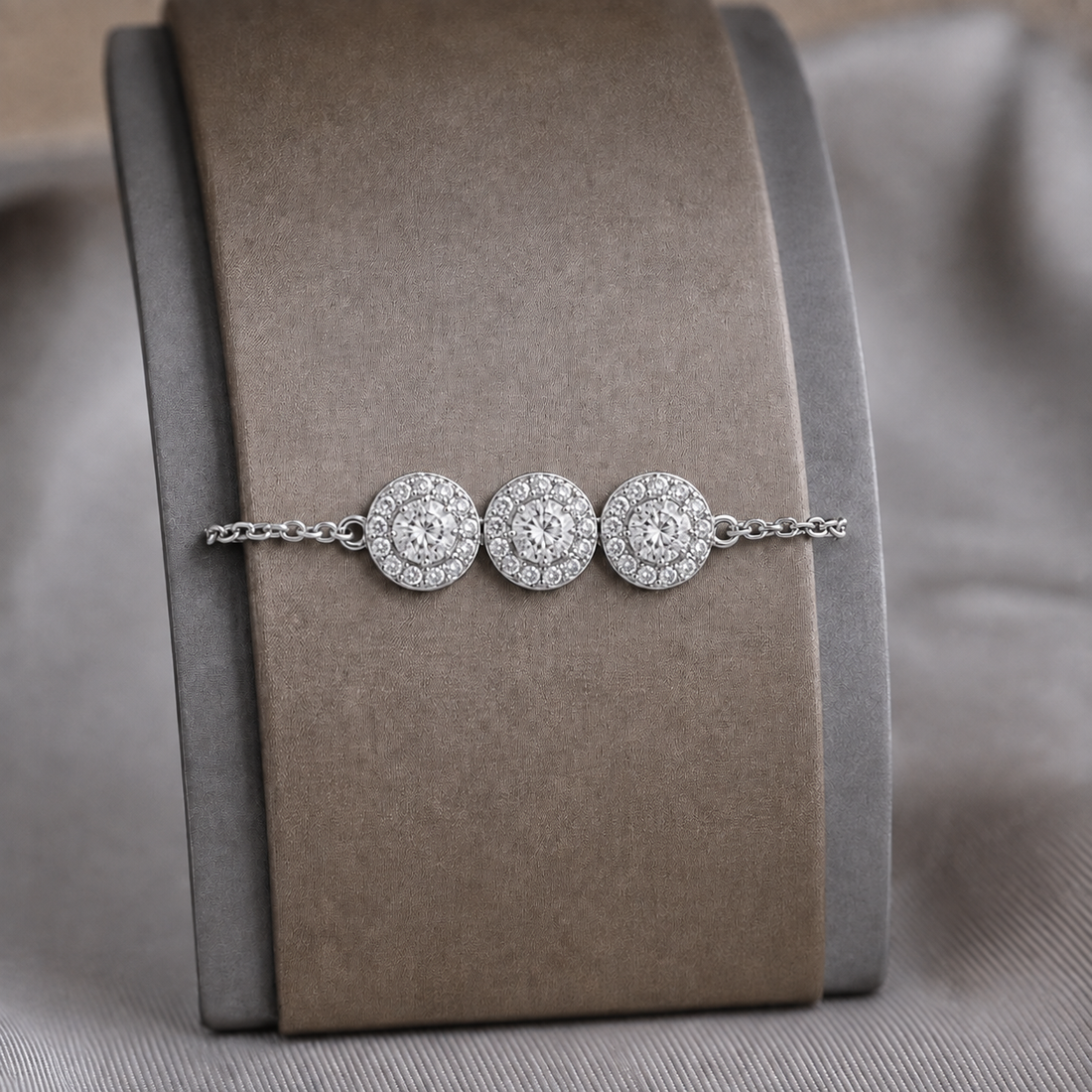 A silver-toned bracelet with three circular pavé medallions, displayed on a brown velvet jewelry stand against a soft gray fabric background.
