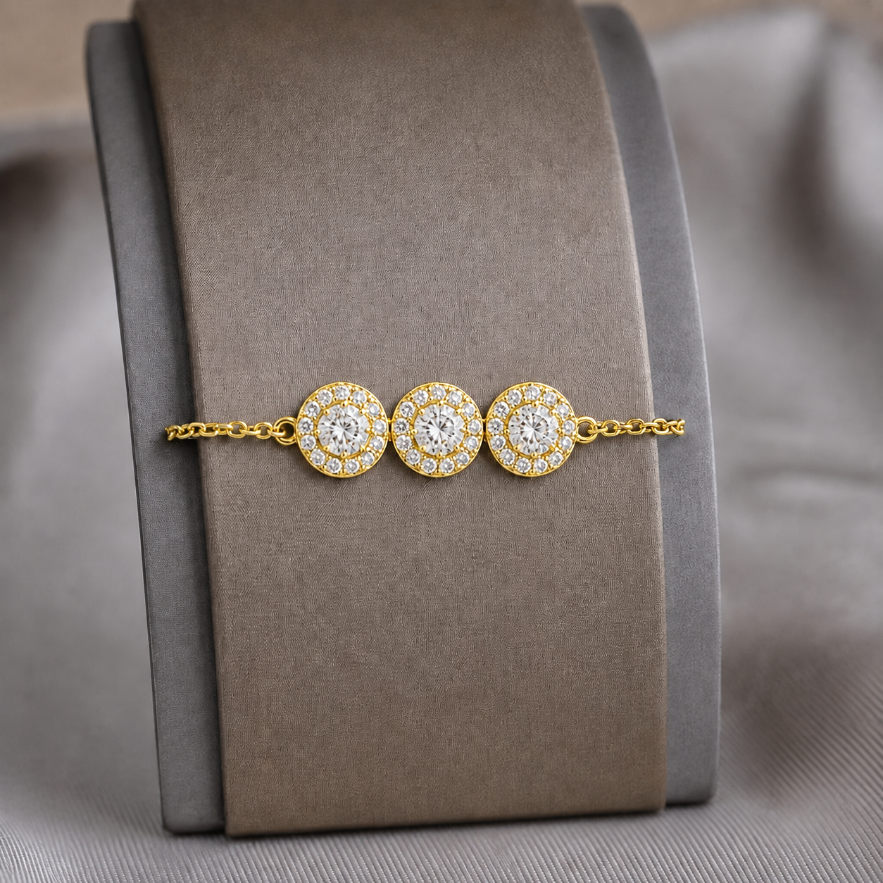 A Gold-toned bracelet with three circular pavé medallions, displayed on a brown velvet jewelry stand against a soft gray fabric background.