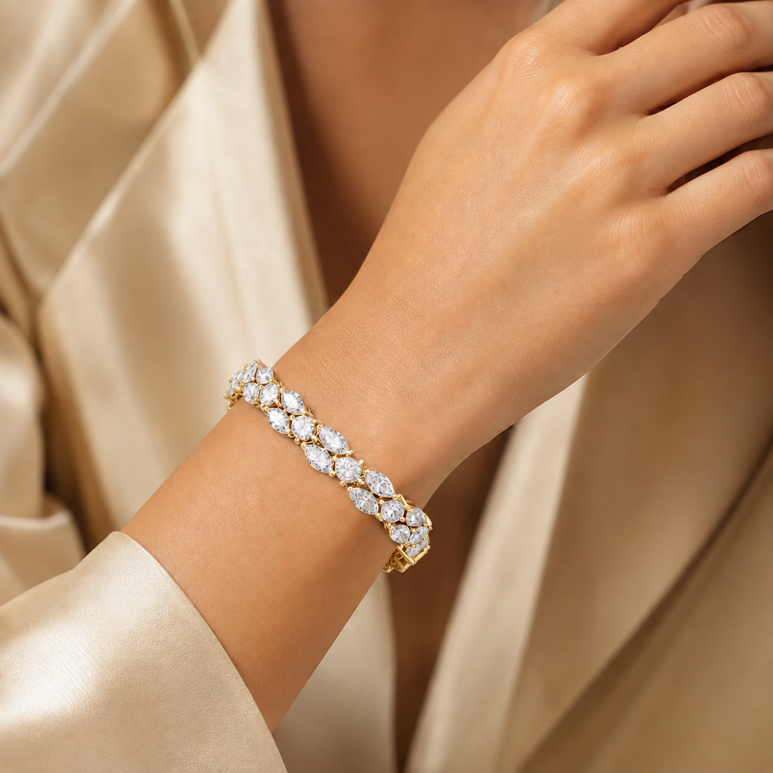 A close-up of a woman's wrist wearing a gold tennis bracelet set with clear marquise-cut gemstones, styled with a cream satin robe.